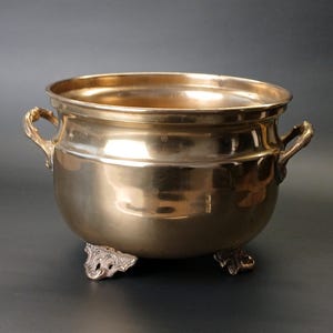 Puede incluir: Un cuenco de latón pulido con un cuerpo liso y redondeado y una abertura ancha. El recipiente cuenta con dos asas ornamentadas y texturizadas en los lados opuestos y se apoya sobre tres pies decorativos con volutas que elevan la base.