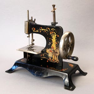 May include: A vintage black metal toy sewing machine featuring a hand crank, a silver-toned wheel, and a needle assembly. The body is decorated with gold-toned floral scrollwork and a colorful illustration of a girl standing next to a wolf.