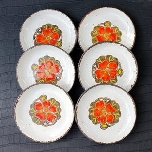 Op de afbeelding: Set van zes witte keramische borden met een levendig oranje bloemmotief in het midden. De bloemen zijn omgeven door een bruine en groene rand. De borden hebben een gespikkelde textuur en een bruine rand.