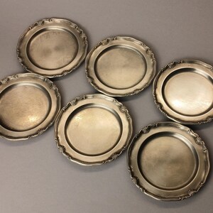 Puede incluir: Seis platos antiguos de tono plateado con bordes festoneados ornamentados. Los platos tienen un centro ligeramente c&oacute;ncavo y un borde decorativo. Est&aacute;n dispuestos sobre una superficie gris, mostrando su calidad reflectante y encanto vintage.