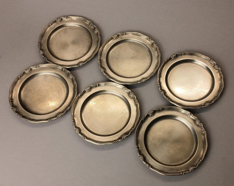 Puede incluir: Seis platos antiguos de tono plateado con bordes festoneados ornamentados. Los platos tienen un centro ligeramente c&oacute;ncavo y un borde decorativo. Est&aacute;n dispuestos sobre una superficie gris, mostrando su calidad reflectante y encanto vintage.