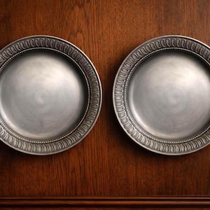 May include: Two circular pewter-colored plates featuring a smooth, matte center and a decorative rim with a repeating vertical ribbed pattern and a beaded inner border. The plates have a metallic finish and are positioned side by side.