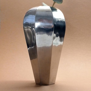 Può includere: Un vaso alto color argento con un design geometrico sfaccettato. Il vaso ha una base stretta che si allarga verso l'alto, con un piccolo rametto di verde che si estende dall'apertura. Il vaso è in metallo riflettente.