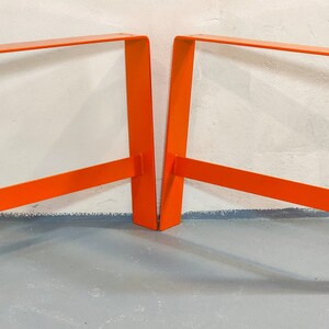 May include: Two bright orange metal table legs, each with a rectangular frame and a single crossbar. The legs are standing on a gray concrete floor.