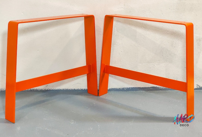 May include: Two bright orange metal table legs, each with a rectangular frame and a single crossbar. The legs are standing on a gray concrete floor.