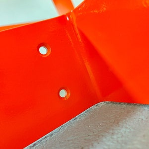 May include: Close-up of a bright orange metal corner bracket with two holes drilled into it. The bracket is painted a glossy orange.