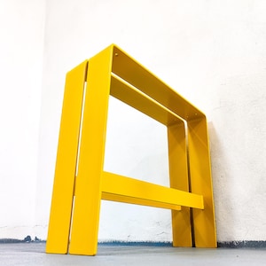 Könnte beinhalten: Ein hellgelber Metallhocker mit minimalistischem Design. Der Hocker hat einen rechteckigen Rahmen mit zwei horizontalen Stangen und zwei vertikalen Beinen. Der Hocker ist in einer einfarbigen, leuchtenden gelben Farbe lackiert. Der Hocker ist für den Innen- und Außenbereich geeignet.