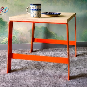 Puede incluir: Una mesa pequeña, cuadrada, de madera con patas de metal naranja. La mesa está sobre una alfombra gris. Una taza de cerámica azul y blanca y un platillo azul y blanco están sobre la mesa.