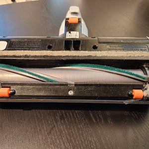 May include: Close-up of a vacuum cleaner brush head with a green and gray roller brush. The brush head is made of black and white plastic and has orange release buttons on the sides. The brush head is designed for cleaning carpets and upholstery.