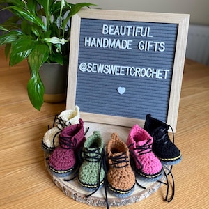 May include: A display of handmade crochet boots in a range of colours: cream, burgundy, green, brown, pink, and black. Each boot has black laces and yellow soles. They are arranged on a wooden slice, with a sign saying "BEAUTIFUL HANDMADE GIFTS" and "@SEWSWEETCROCHET".