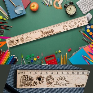 May include: A wooden ruler with a dinosaur, backpack, and globe design. The ruler is 30 cm long and has a personalized name "Jonas" and the text "bereit f&uuml;r das Abenteuer Schule!"  The ruler is on a green background with school supplies.