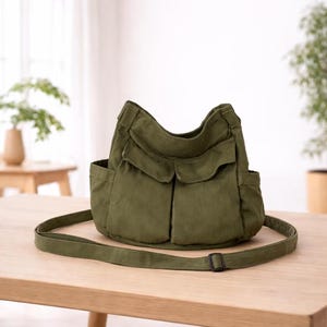 May include: Olive green shoulder bag with multiple exterior pockets and a long, adjustable strap. The bag is made of a durable fabric and sits on a wooden table. The design includes two front flap pockets and two side pockets.