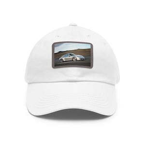 Può includere: Cappellino da baseball bianco con una toppa rettangolare raffigurante un'auto sportiva argentata su una strada. L'auto è il punto focale, sullo sfondo di una strada e di un cielo nuvoloso. Il cappellino è realizzato in materiale morbido e resistente.