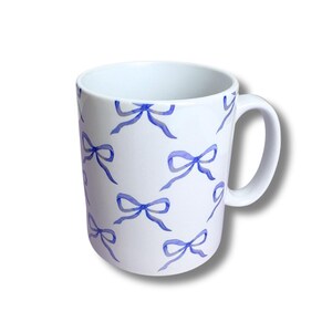May include: White ceramic mug with a repeating pattern of blue watercolor bows. The mug has a curved handle and a simple, clean design. The bows are in various sizes and orientations, adding visual interest to the mug.