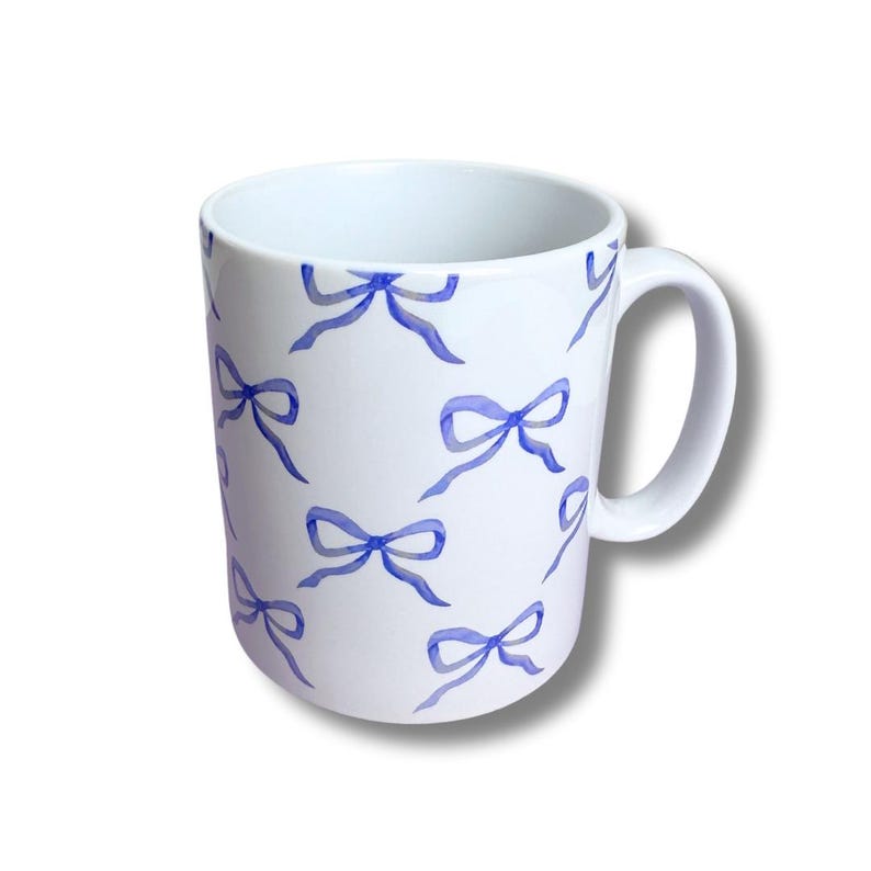 May include: White ceramic mug with a repeating pattern of blue watercolor bows. The mug has a curved handle and a simple, clean design. The bows are in various sizes and orientations, adding visual interest to the mug.