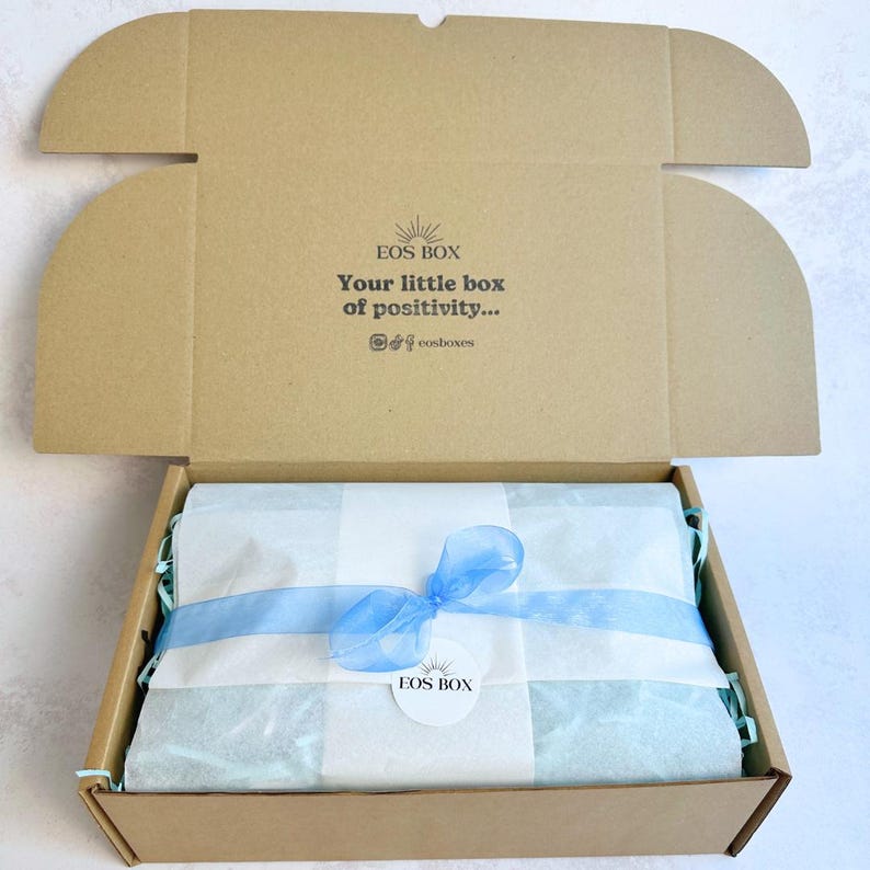 May include: Open cardboard box with a lid, revealing a wrapped gift inside. The gift is wrapped in white and light blue tissue paper and tied with a light blue ribbon. The box has the text "EOS BOX" and "Your little box of positivity..."