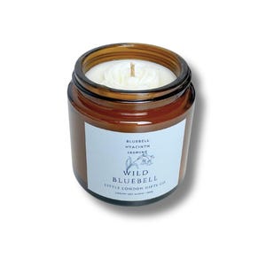 May include: A small, brown glass jar candle with a white label. The label reads "Wild Bluebell" with the scent notes "Bluebell, Hyacinth, Jasmine." The candle has a white wax and a single wick. The jar is filled with a light-colored wax.