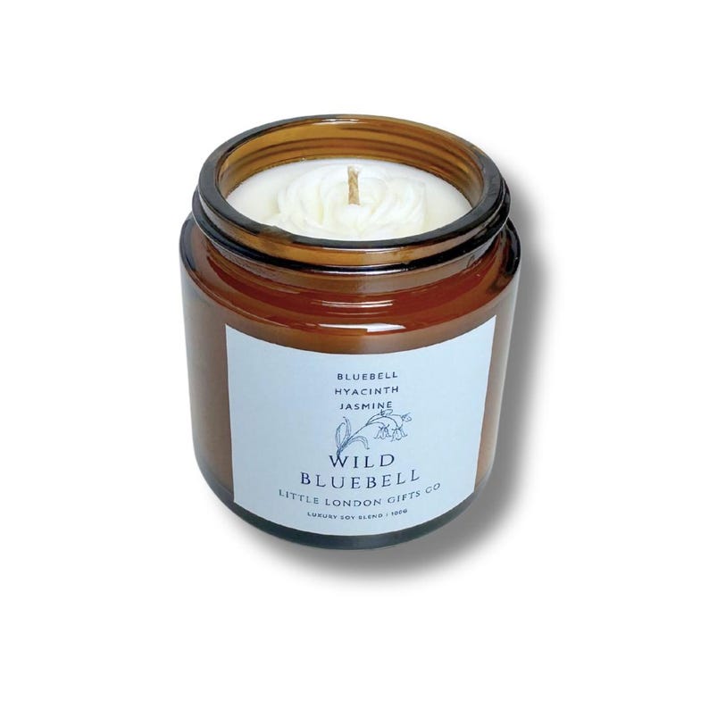 May include: A small, brown glass jar candle with a white label. The label reads "Wild Bluebell" with the scent notes "Bluebell, Hyacinth, Jasmine." The candle has a white wax and a single wick. The jar is filled with a light-colored wax.