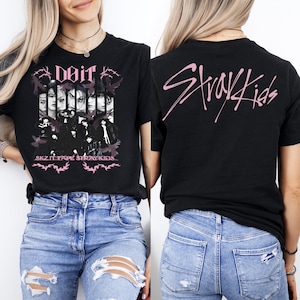 K&ouml;nnte beinhalten: Schwarzes T-Shirt mit einem rosa Grafikdesign mit den Texten "Doit" und "SKZ IT TAPE STRAY KIDS". Auf der R&uuml;ckseite des T-Shirts steht der Text "Stray Kids" in Rosa. Das T-Shirt wird mit einer blauen Jeans getragen.
