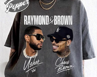 May include: A charcoal gray t-shirt featuring the text RAYMOND & BROWN at the top. Below, two portraits of men wearing sunglasses and gold chains are printed. The left portrait is labeled Usher with a crown icon, and the right is labeled Chris Brown.