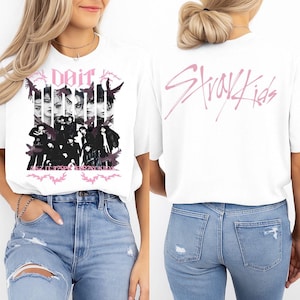 K&ouml;nnte beinhalten: Wei&szlig;es T-Shirt mit einer Schwarz-Wei&szlig;-Grafik einer Personengruppe und dem rosa Text "Doit" und "SKZ IT TAR STRAYKIDS". Auf der R&uuml;ckseite des Shirts steht "Stray Kids" in Rosa.