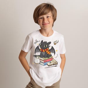 May include: White t-shirt featuring a cartoon dragon with a scarf, sitting on books. The dragon has large eyes and wings. A small owl and other magical elements are also depicted. The shirt is worn by a person.
