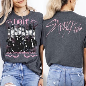 K&ouml;nnte beinhalten: Dunkelgraues T-Shirt mit einem rosa Aufdruck auf der Vorderseite, einschlie&szlig;lich der Texte "Doit" und "SKZ IT TAPE STRAY KIDS". Auf der R&uuml;ckseite des T-Shirts steht der rosa Text "Stray Kids".