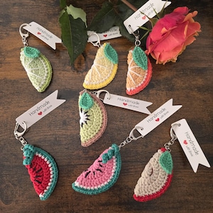 May include: A collection of handmade crochet fruit slice keychains, including lemon, orange, kiwi, watermelon, and apple designs. Each keychain features a silver ring and a tag that reads "Handmade with love."