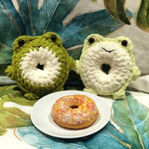 May include: Two green crocheted frog plushies with white donut-shaped centres, sitting on a white surface with a glazed donut with colourful sprinkles.
