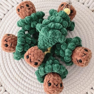 May include: A collection of handmade crochet Christmas tree keychains. Each keychain features a green tree with a brown, smiling face ornament. The keychains are made of soft, plush yarn and are designed to be decorative.