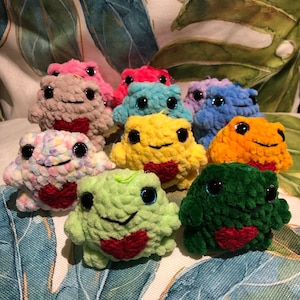 May include: A collection of ten crocheted frog plushies in various colours, including pink, blue, yellow, green, and orange. Each frog has a red heart on its chest.