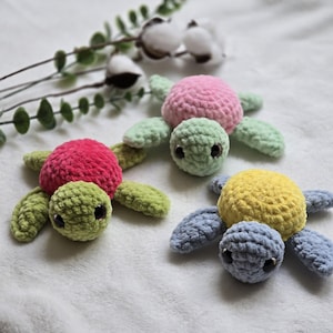 Puede incluir: Tres tortugas hechas a mano de ganchillo en tonos rosa, verde, amarillo y azul. Cada tortuga tiene un caparazón redondeado y extremidades en forma de aleta. Se exhiben sobre una superficie blanca con ramitas de vegetación y algodón.