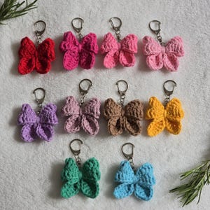 May include: Twelve crocheted bow keychains in various colors, including pink, purple, green, blue, yellow, and brown. Each keychain features a silver metal ring.