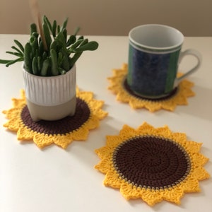 Könnte beinhalten: Drei gehäkelte Sonnenblumen-Untersetzer in Gelb und Braun. Die Untersetzer liegen auf einer weißen Oberfläche mit einer kleinen Topfpflanze im Hintergrund.