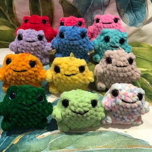 Può includere: Una collezione di 11 giocattoli di rana di peluche all'uncinetto in vari colori, tra cui rosso, viola, arancione, giallo, verde, blu, rosa e grigio. Le rane hanno grandi occhi neri e un'espressione sorridente.