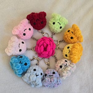 May include: A collection of 12 crocheted plush keychains in various colors, including pink, blue, green, yellow, and red. Each keychain features a cute, smiling face and is attached to a silver metal ring.
