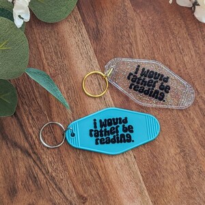 Puede incluir: Dos llaveros con el texto "I would rather be reading." Uno es transparente con purpurina, el otro turquesa. Ambos tienen llaveros de metal. Los llaveros están sobre una superficie de madera con detalles florales.