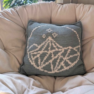 Puede incluir: Una almohada de crochet azul claro con un diseño geométrico blanco en el centro. La almohada está sentada en una silla blanca.