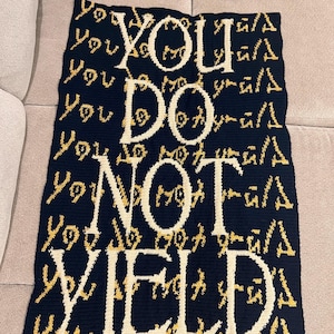 Puede incluir: Una manta de crochet negra y dorada con las palabras "YOU DO NOT YIELD" en blanco.
