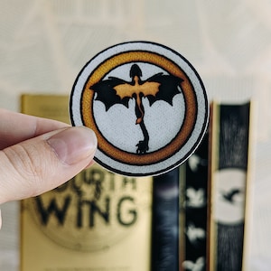 Könnte beinhalten: Ein runder Aufkleber mit einem Drachendesign in einem goldenen Ring. Der Drache hat schwarze Flügel und einen goldenen Körper. Der Aufkleber wird vor einem Buch mit dem Titel "Fourth Wing" in goldener Schrift gehalten.
