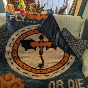 K&ouml;nnte beinhalten: Eine blaue und orangefarbene geh&auml;kelte Decke mit einem Drachenmotiv und dem Text "FLY... OR DIE".