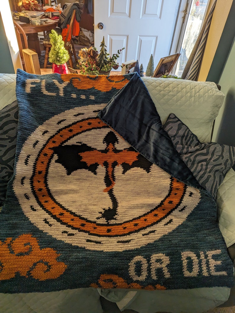 K&ouml;nnte beinhalten: Eine blaue und orangefarbene geh&auml;kelte Decke mit einem Drachenmotiv und dem Text "FLY... OR DIE".