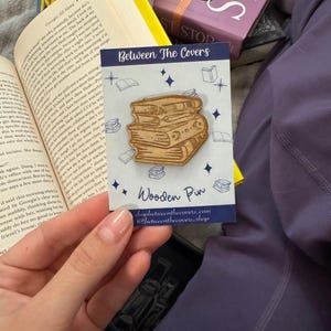 Pode incluir: Um alfinete de madeira com uma pilha de livros. O alfinete é castanho claro e tem um design detalhado. O alfinete está num cartão com o texto "Between The Covers" e "Wooden Pin".
