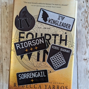 Op de afbeelding: Een boek getiteld "Fourth Wing" met verschillende zwart-witte patches op de cover. De patches hebben tekst als "IV WINGLEADER", "RIORSON" en "SORRENGAIL". Het boek heeft een gouden kaft met de naam van de auteur, Rebecca Yarros.