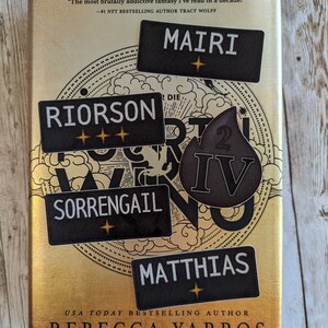 Op de afbeelding: Een boek getiteld "Fourth Wing" van Rebecca Yarros, met zwarte naamlabels met de namen "Mairi", "Riorosn", "Sorrengail" en "Matthias". De cover is goudkleurig met een cirkelvormig ontwerp en de tekst "The most brutally addictive fantasy I've read in a decade!"