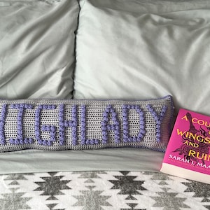 Puede incluir: Una almohada de crochet morada y gris con la palabra "HIGH LADY" escrita en hilo morado. Un libro rosa titulado "A Court of Wings and Ruin" de Sarah J. Maas está sobre una cama con una manta de patrón gris y blanco.