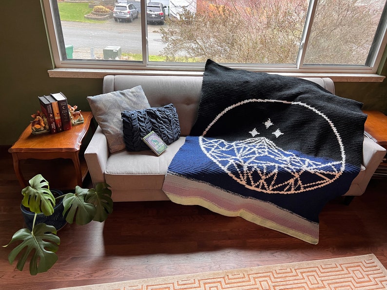 May include: A crocheted blanket with a geometric mountain design and three stars in a circle on a black background. The blanket is draped over a light gray couch with pillows.