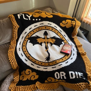 K&ouml;nnte beinhalten: Eine schwarz-wei&szlig;e H&auml;keldecke mit gelbem Rand. Die Decke zeigt einen Drachen in einem Kreis mit den Worten "FLY... OR DIE" und einem Wolkenmuster. Ein DVD-Geh&auml;use f&uuml;r den Film "Drachenz&auml;hmen leicht gemacht" liegt auf der Decke.