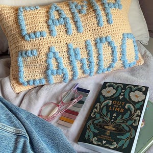 May include: A yellow crocheted pillow with blue pom-poms spelling out "CAMP CHINO" on a bed with a book titled "Out on a Limb" by Hannah Bonham-Young.
