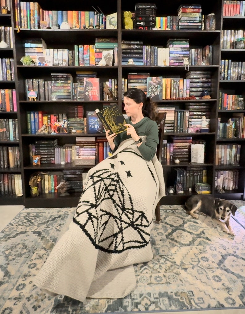 May include: A woman sits in a chair reading a book titled "A Court of Mist and Fury" by Sarah J. Maas. She is wrapped in a beige crochet blanket with a black geometric design. A small dog sits on the floor next to her.